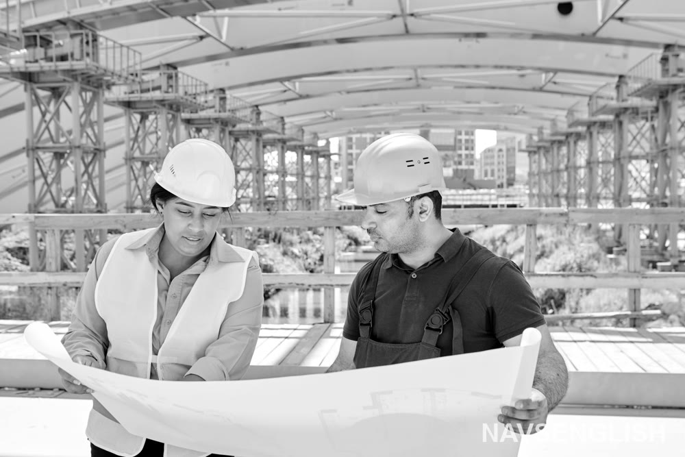 Công trình xây dựng, phía sau có cấu trúc được che một phần và giàn giáo. Bên trái một người phụ nữ đội nón bảo hộ, áo sáng màu, mặc vest an toàn, đang cầm một tài liệu lớn và nhìn vào nó. Bên phải một người đàn ông đội nón bảo hộ, áo tối màu, mặc yếm, cũng đang cầm tài liệu và xem xét nó. Tài liệu được trải ra giữa hai người. Phía sau có cây xanh và vài toà nhà ở xa